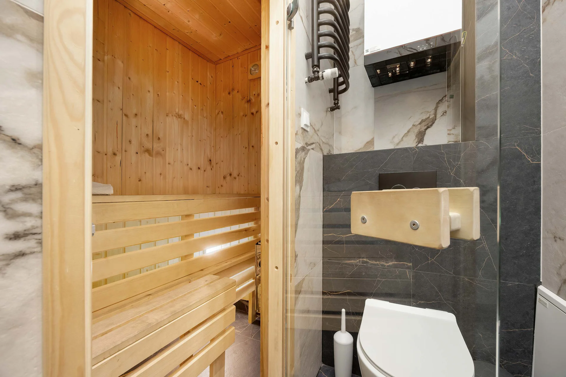 A modern bathroom featuring a glass shower, a toilet, and a compact wooden sauna. The combination of stone tiles and natural wood creates a luxurious, spa-like atmosphere.