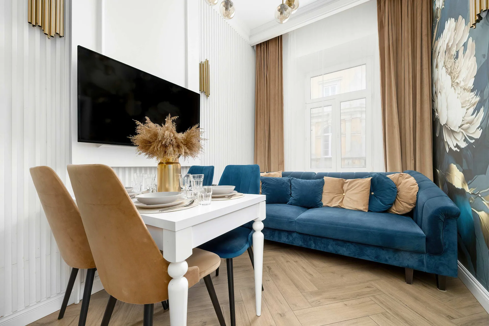 A bright and elegant living room with a comfortable blue sofa and a dining table surrounded by beige chairs. The large windows and decorative wallpaper create a cozy and welcoming atmosphere.