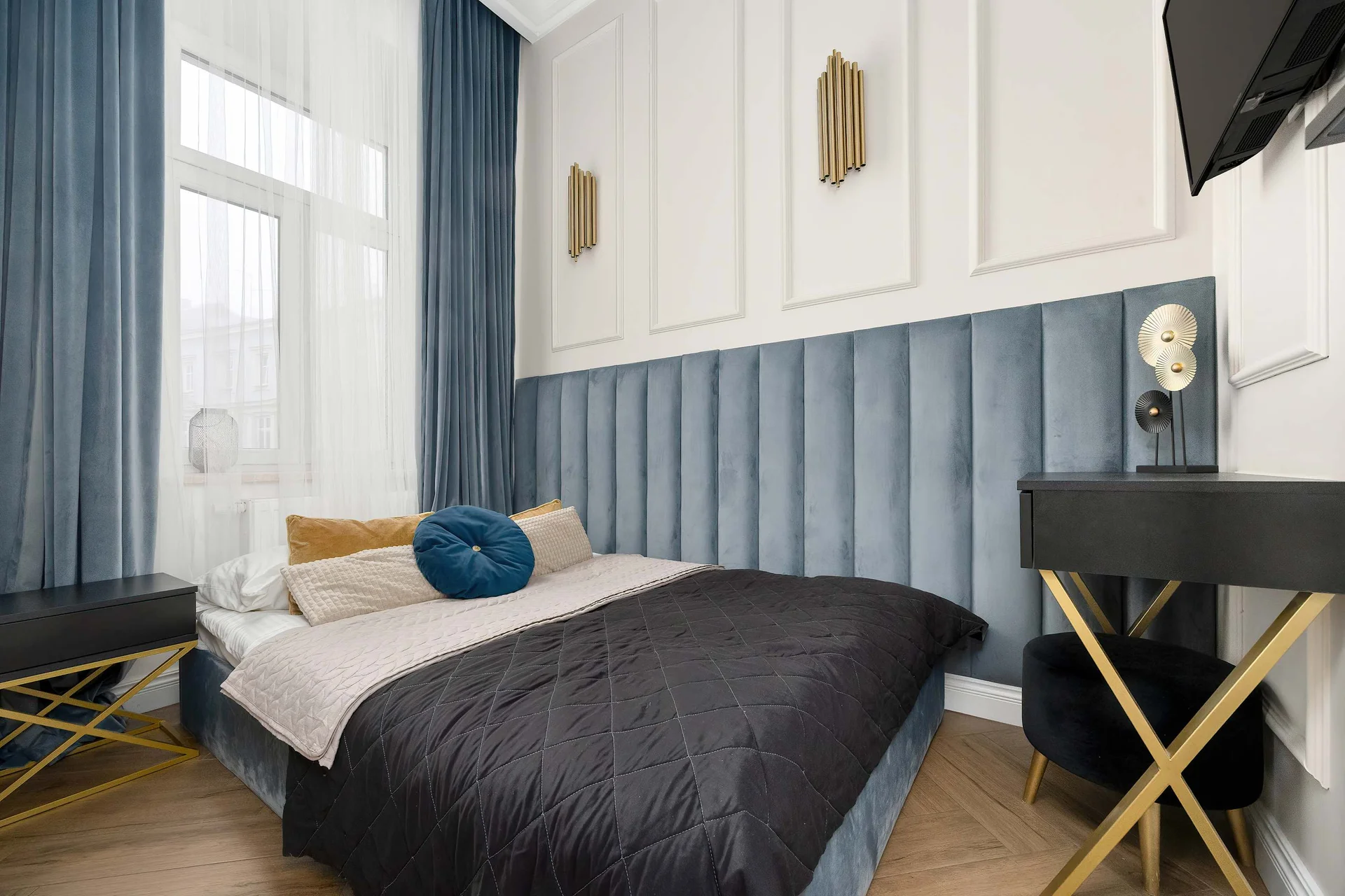 A comfortable bedroom with a large double bed, dark bedding, and blue upholstered wall panels. The large window allows plenty of natural light to enter, creating a peaceful space to relax.