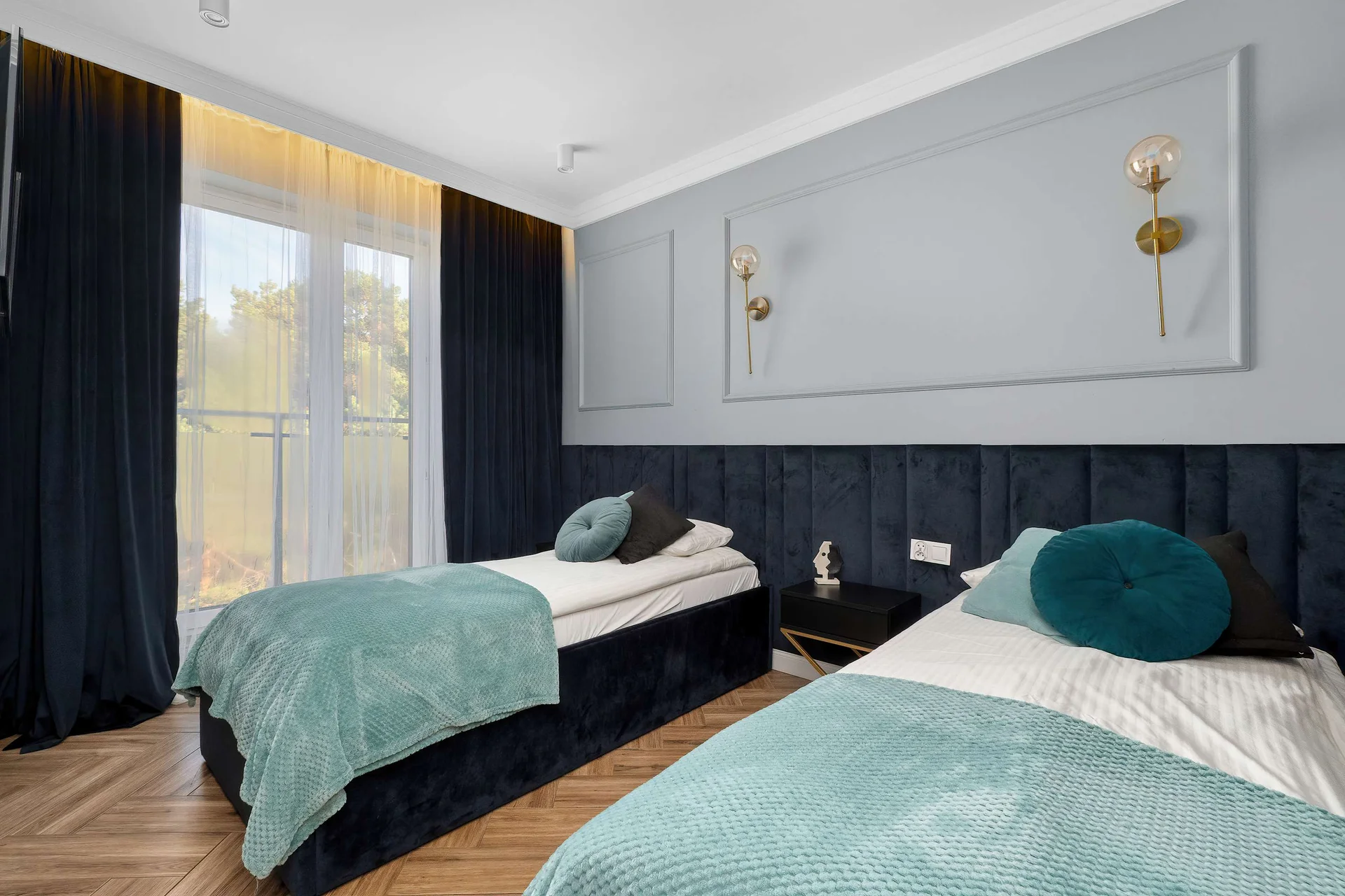 Modern bedroom with two single beds, teal bedding, dark accent wall and gold pendant lights.