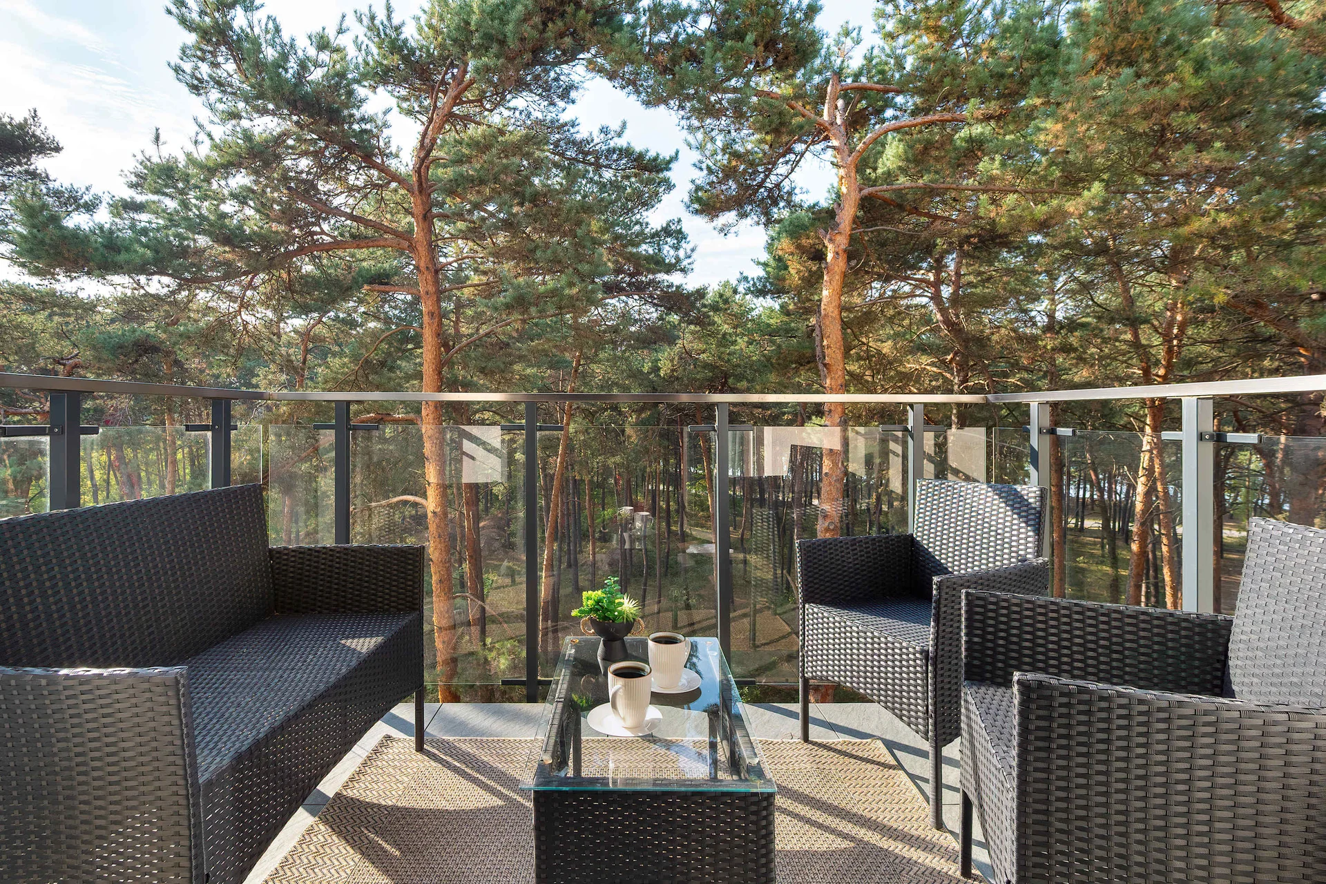 Spacious balcony with outdoor lounge furniture, decorative plants, and views of surrounding pine trees.