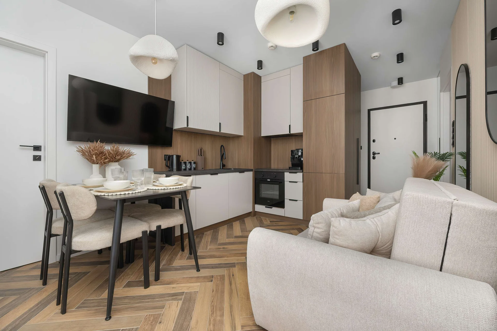 An open-plan living area combining a fully equipped kitchenette, a dining table, and a comfortable sofa. Modern cabinetry, warm wood accents, and large windows create a welcoming space.