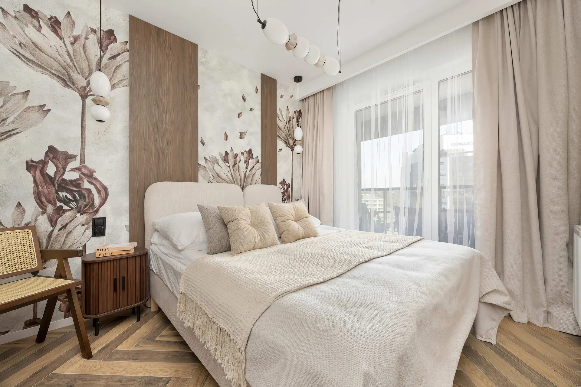 A bright bedroom featuring a comfortable double bed with soft white bedding and decorative cushions. Floral wall art, warm wooden flooring, and neutral furnishings create a relaxing atmosphere.