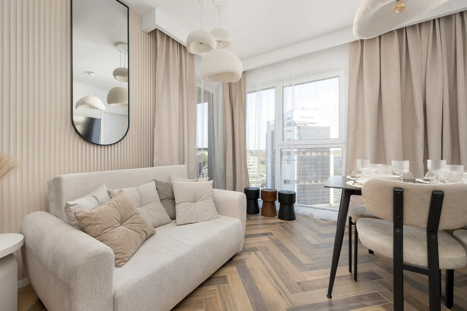 A cozy living room with a comfortable beige sofa, a round dining table, and elegant neutral-toned decor. Large windows with floor-length curtains fill the space with natural light.