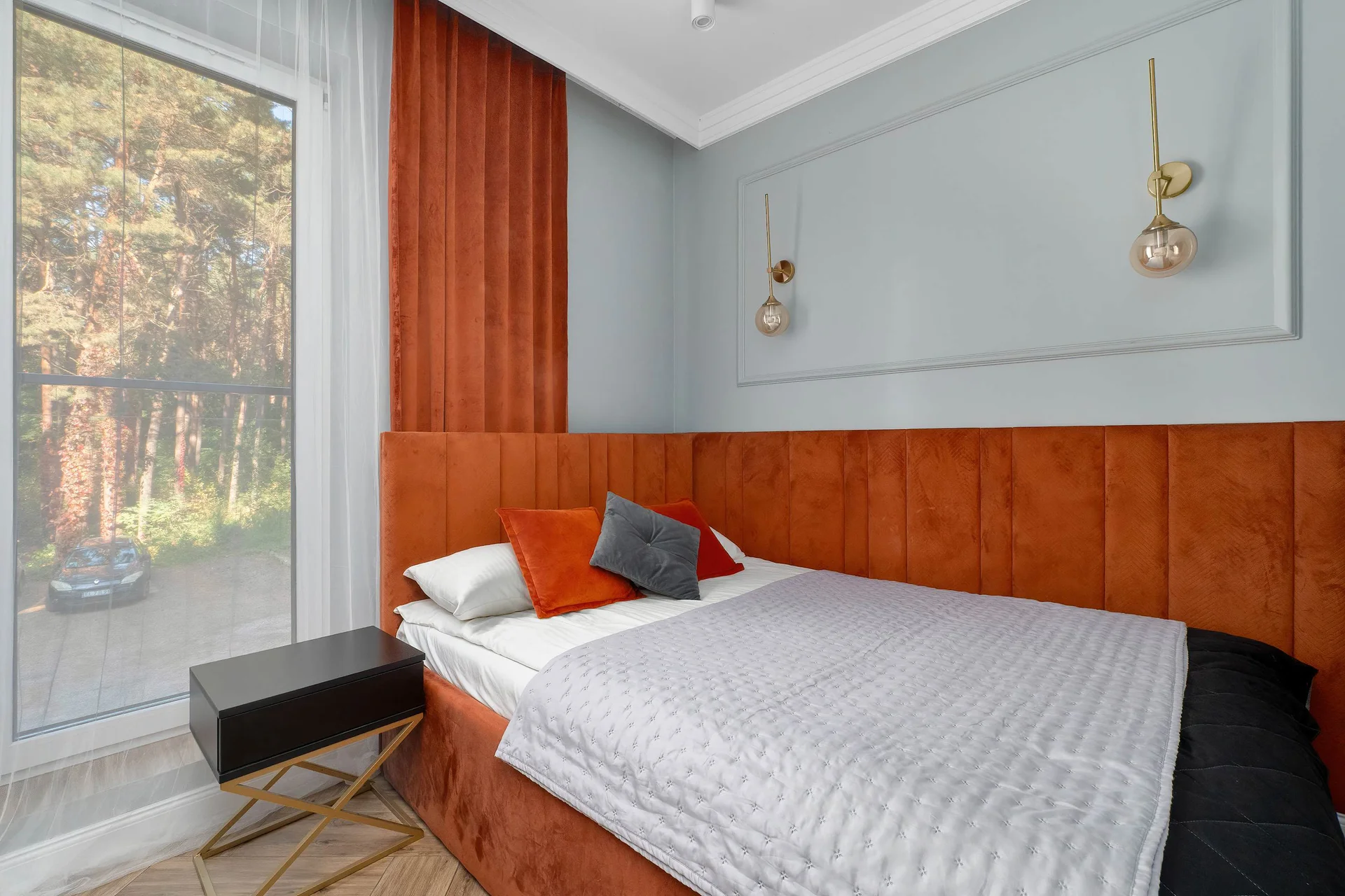 A cozy bedroom contains a double bed with white bedding and an orange accent pillow. A warm wood-paneled wall is visible behind the bed next to a large window showing trees.