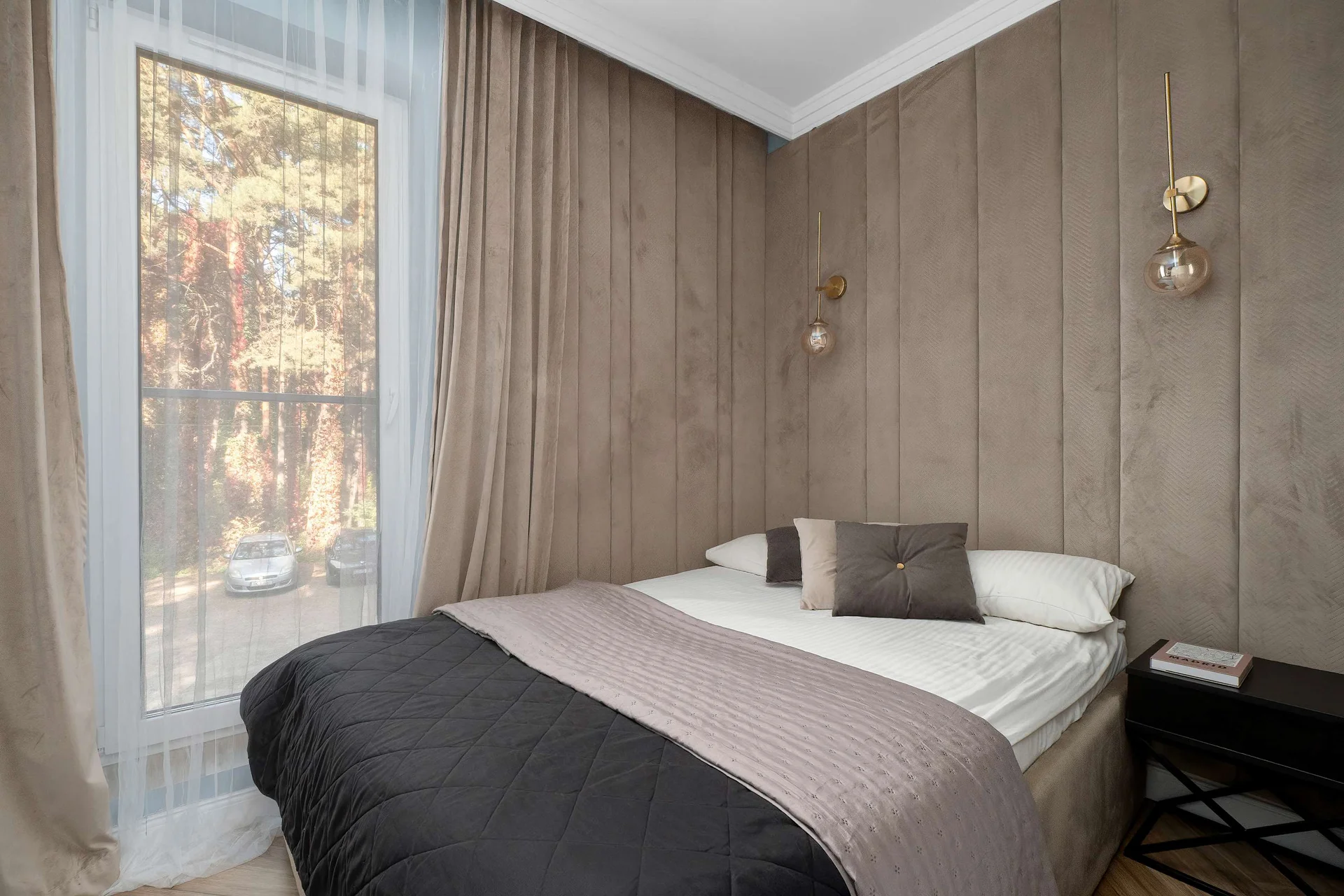 A relaxing second bedroom space offers a double bed, dark gray bedding, and a beige textured wall. A window on the left looks out onto a serene forest.