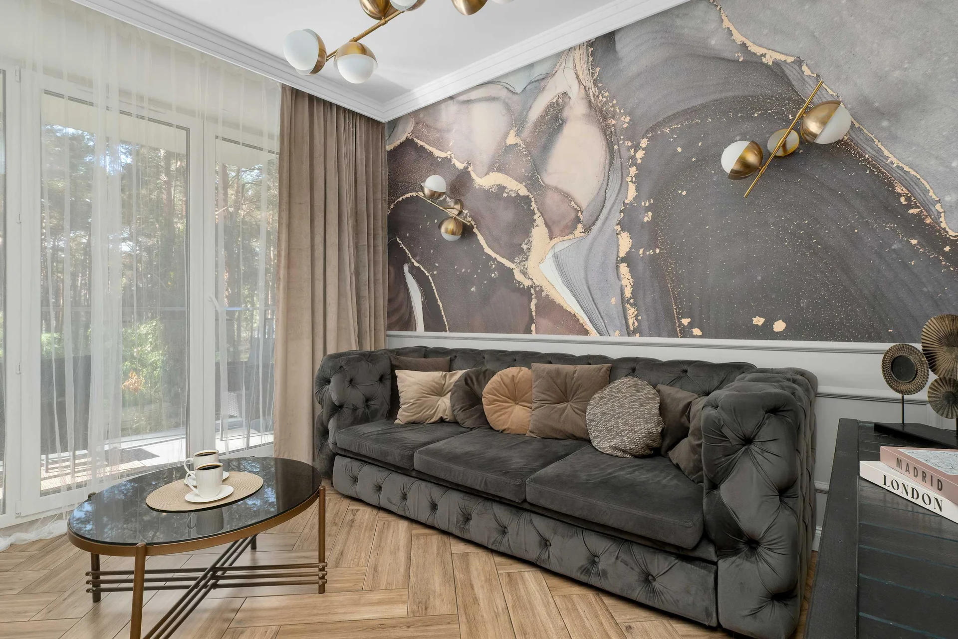 A comfortable living room features a dark tufted sofa, a round coffee table, and an abstract patterned wall. Large windows let in plenty of natural light.