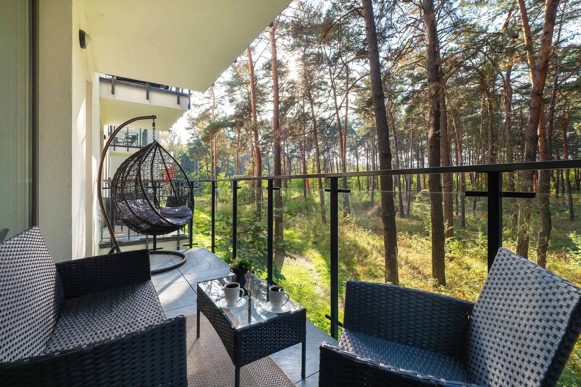 A balcony is equipped with a hanging egg chair and black wicker patio furniture. It overlooks a dense, peaceful pine forest.