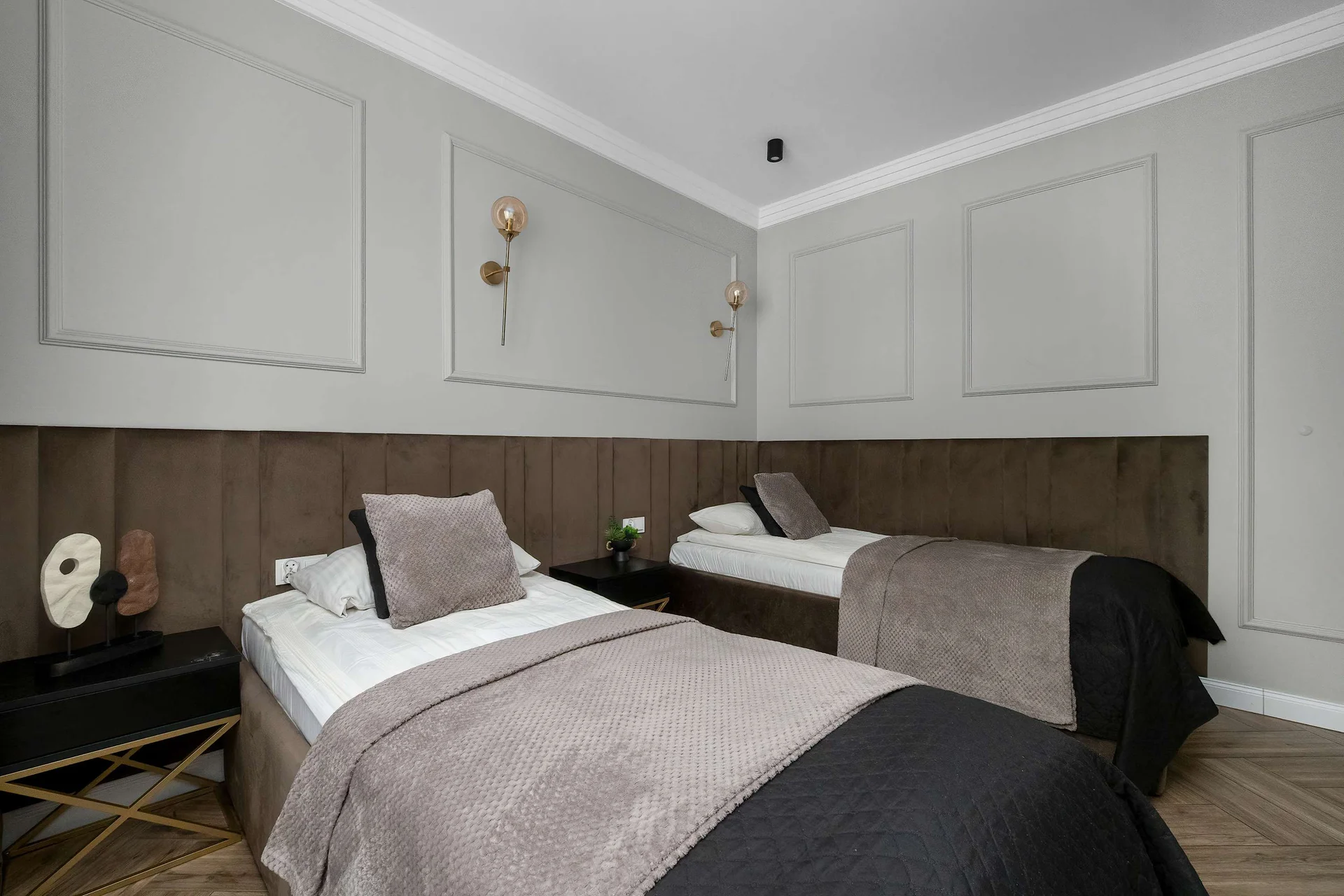 A second bedroom offers two twin beds set against elegant light grey paneled walls. Each bed features a dark, tufted headboard and matching dark throw blankets.
