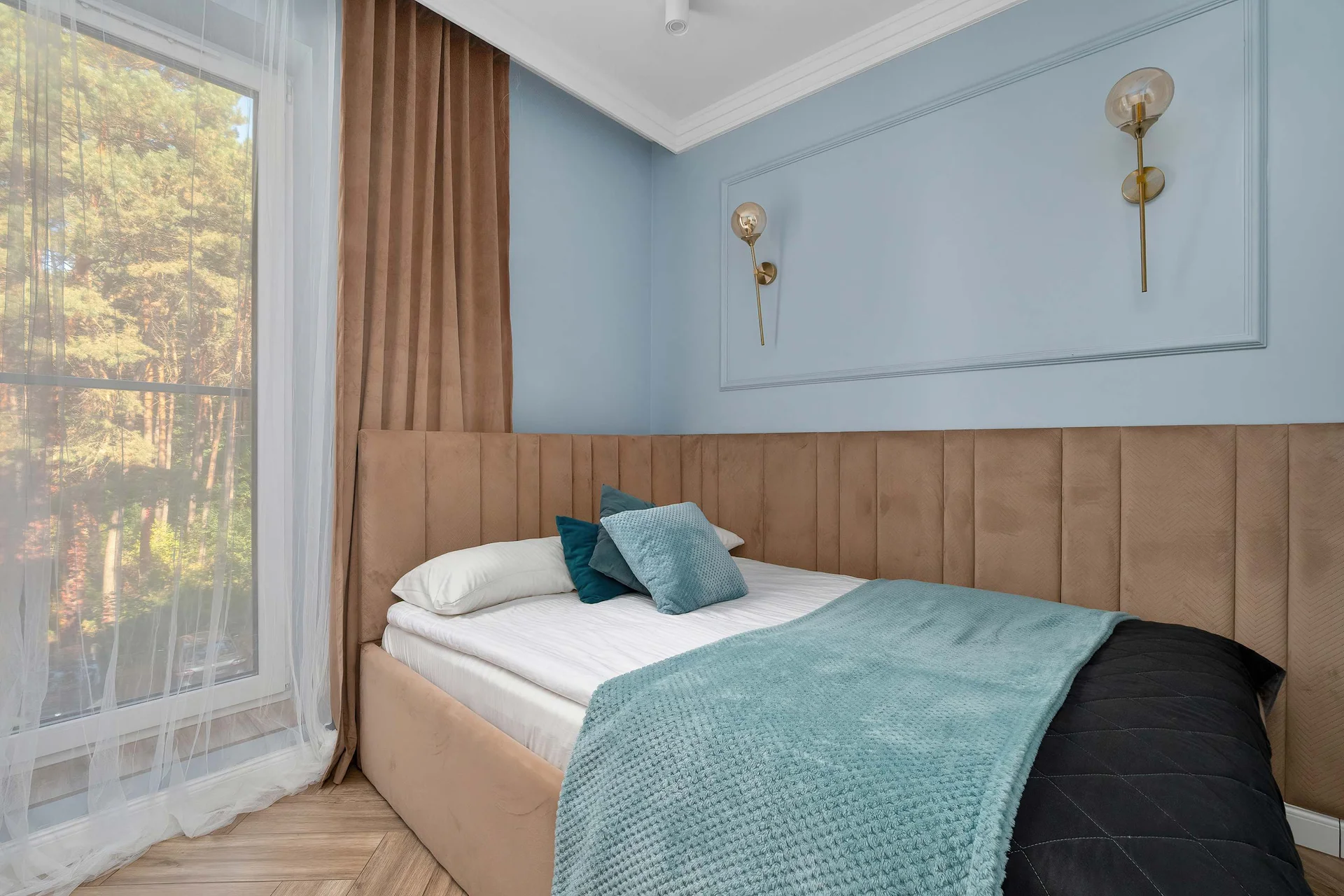 A bright third bedroom showcases light blue walls and a warm wooden paneled headboard. A bright teal blanket adds a pop of color to the bed, while a large window lets in ample natural light.