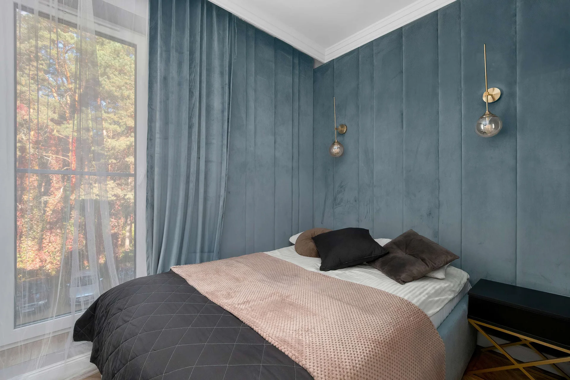 A cozy bedroom is decorated with deep blue walls and floor-to-ceiling curtains framing a large window. A neatly made bed with a dark patterned throw sits at the center of the space.