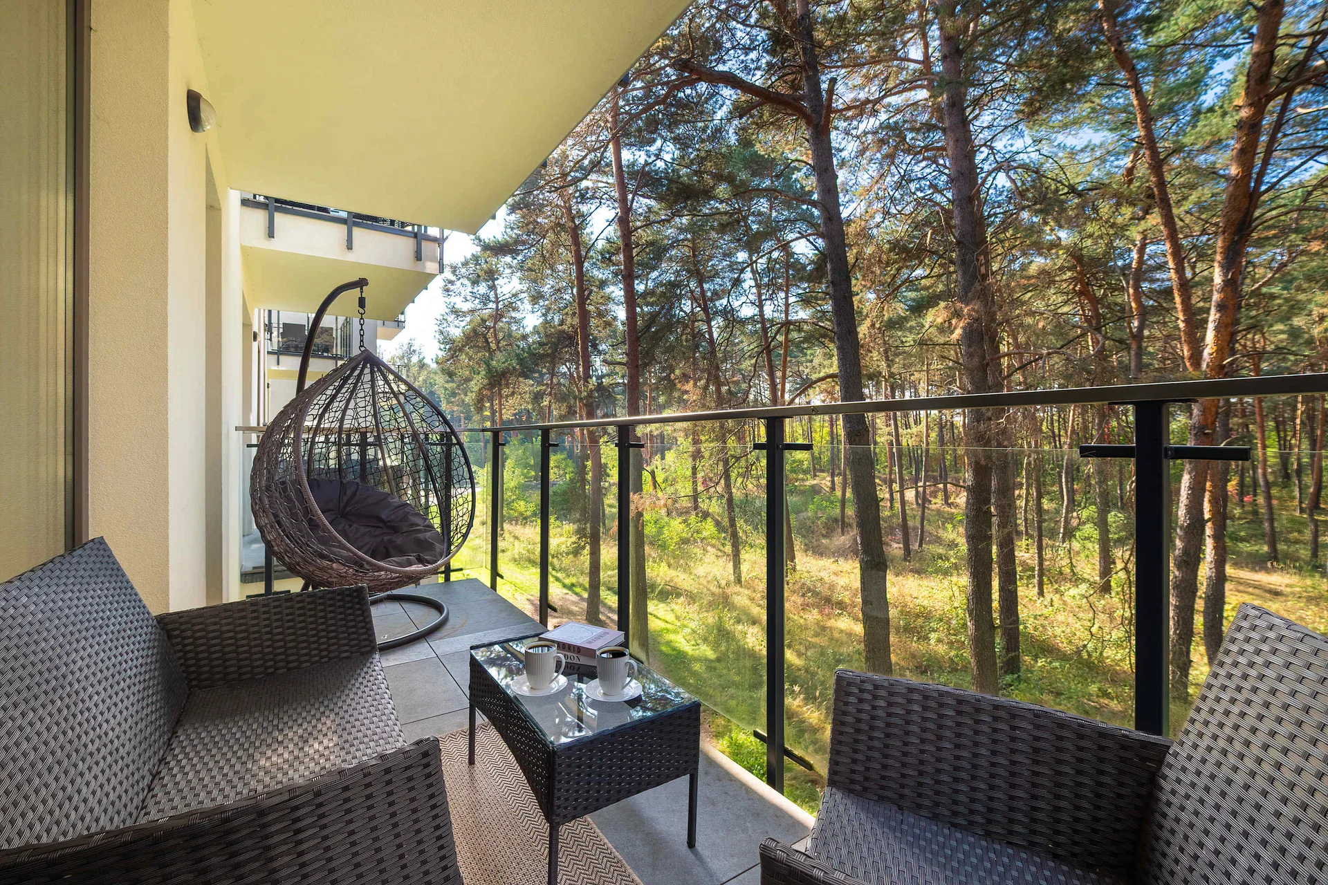 A terrace equipped with comfortable wicker seating and a hanging egg chair provides a relaxing outdoor space. The outdoor area directly overlooks a serene, sunlit pine forest.
