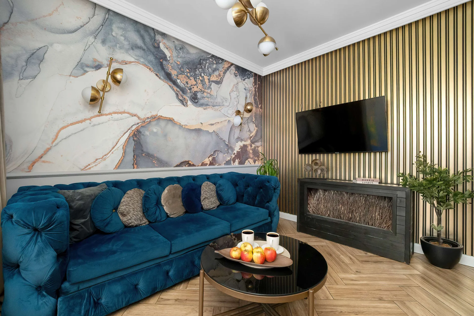 A stylish living room features a plush blue velvet sofa positioned in front of a striking marble-patterned wall mural. A modern flat-screen television is mounted on a textured, slatted wood panel nearby.