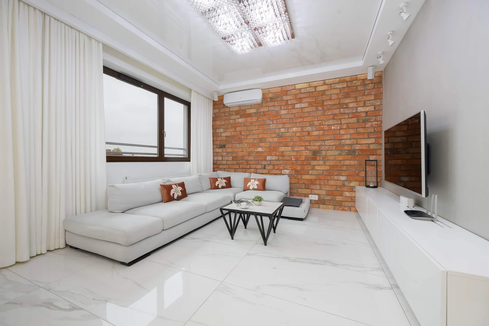 Bright and spacious living room with exposed brick walls, large windows, white furniture, a dining table, and a modern coffee table.