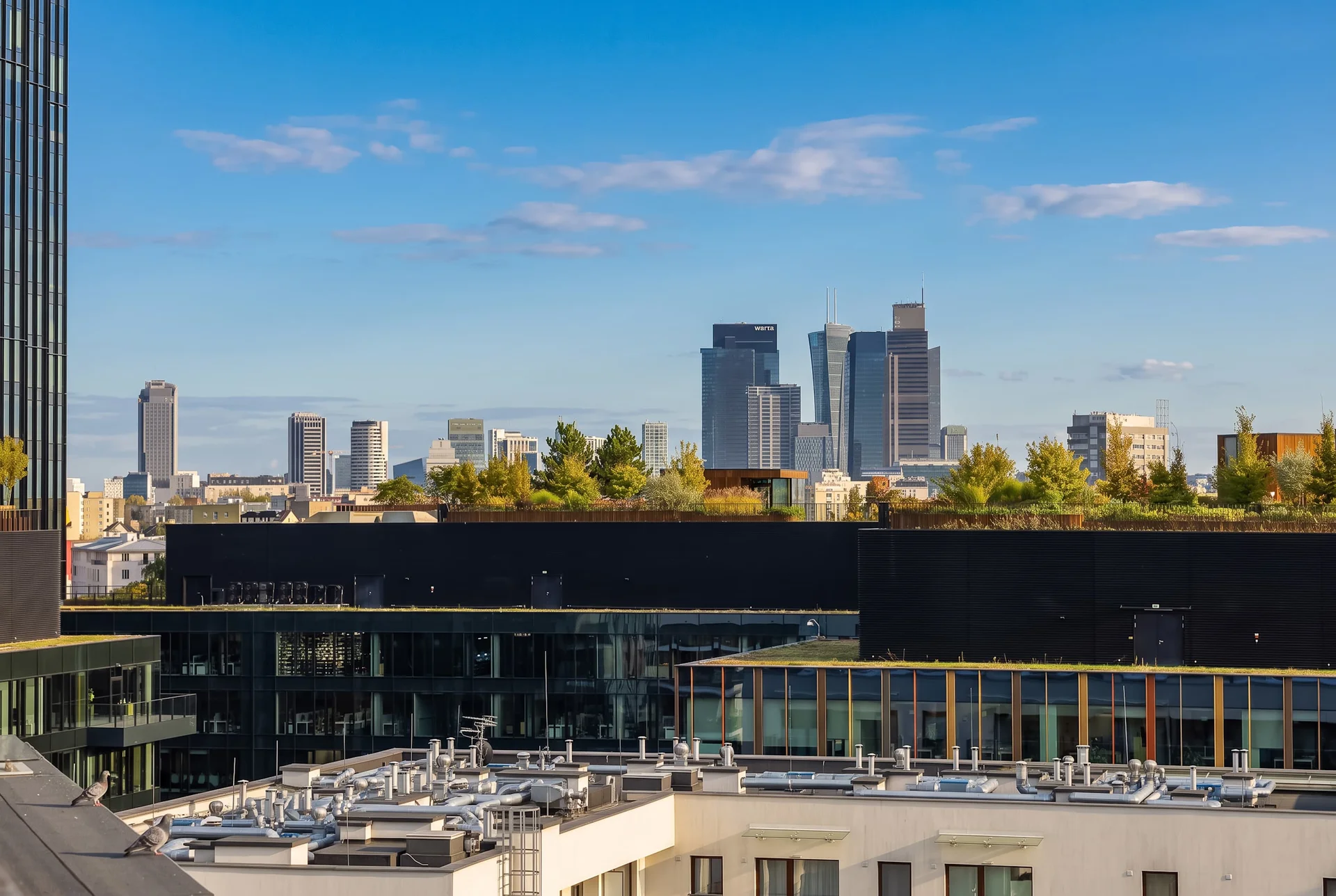 City view from a balcony overlooking modern high-rise buildings and rooftops under a clear blue sky.