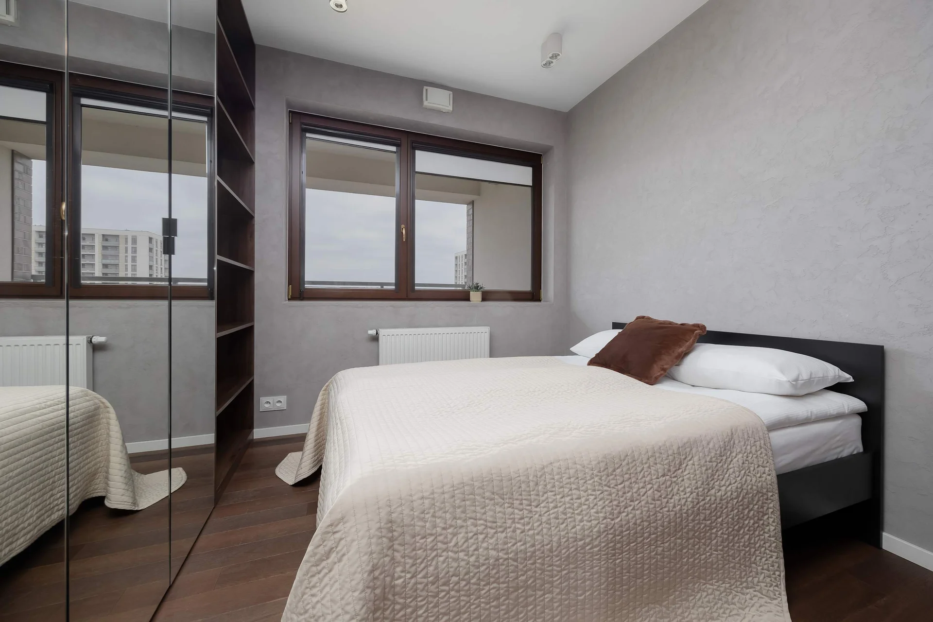 Cozy bedroom with a double bed, beige bedding, dark wooden flooring, large windows, and a built-in wardrobe.