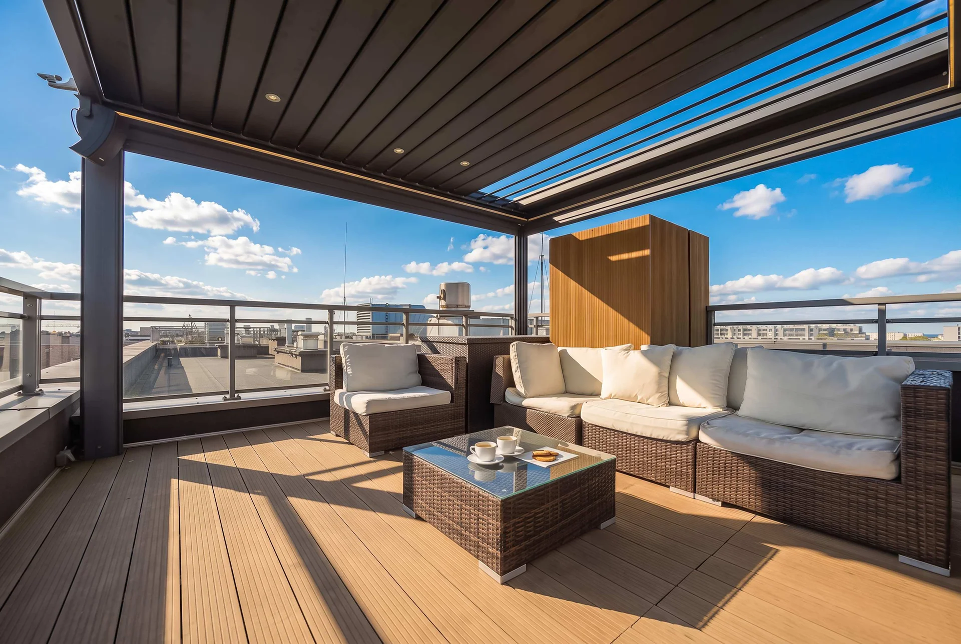 Spacious terrace with outdoor sofas, a coffee table, wooden decking, and a covered pergola overlooking the city.
