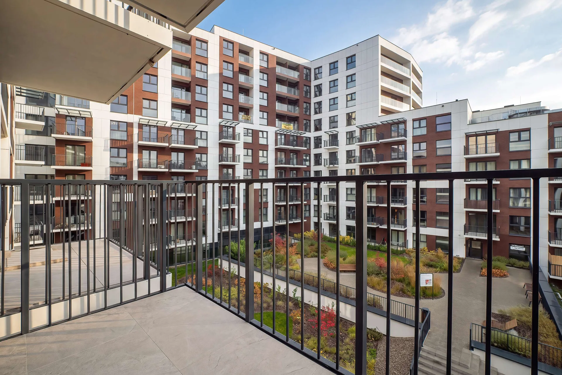 A balcony with black railings overlooking modern residential buildings and a landscaped courtyard.