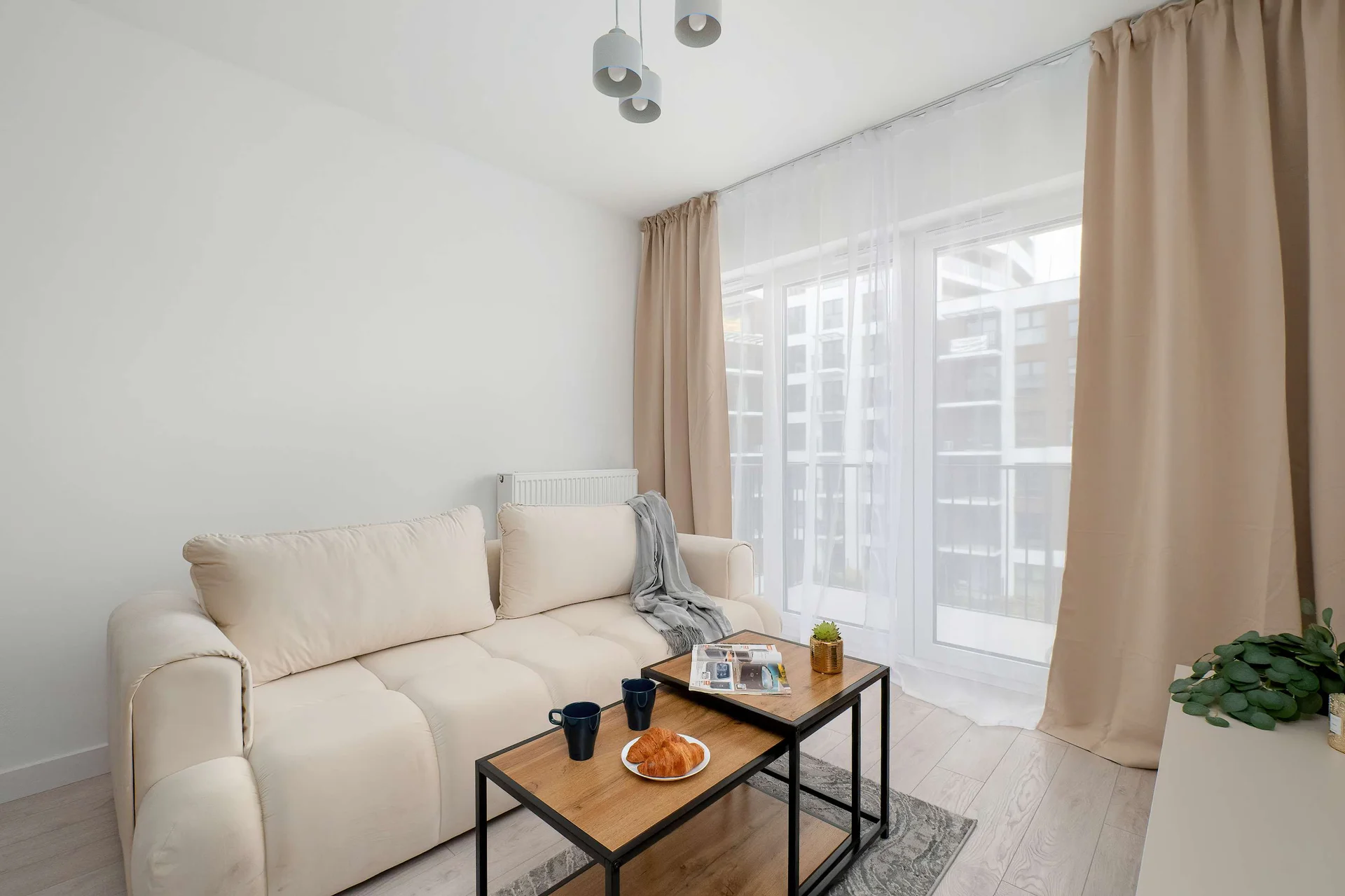 A cozy living room with a cream sofa, a glass coffee table, and large windows with beige curtains.
