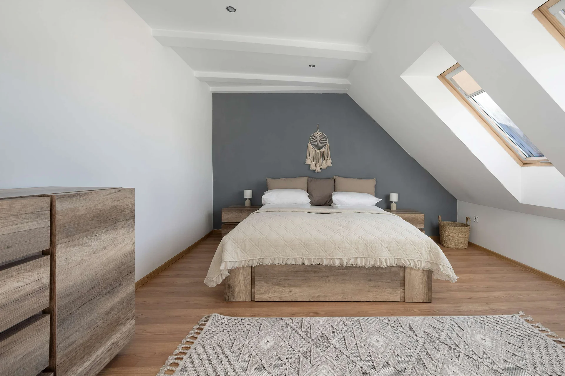A cozy bedroom is situated under a slanted ceiling with a large skylight. A double bed with neutral bedding rests gracefully against a dark grey accent wall.