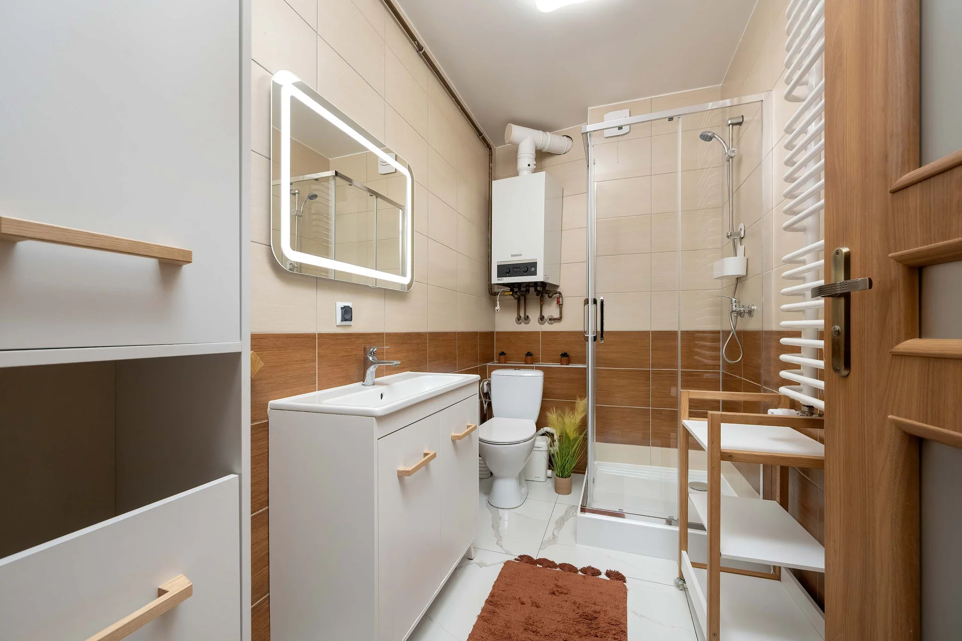 A functional bathroom includes a glass-enclosed shower stall, a white vanity, and a toilet. A white water heater is securely mounted on the wall directly above the toilet area.