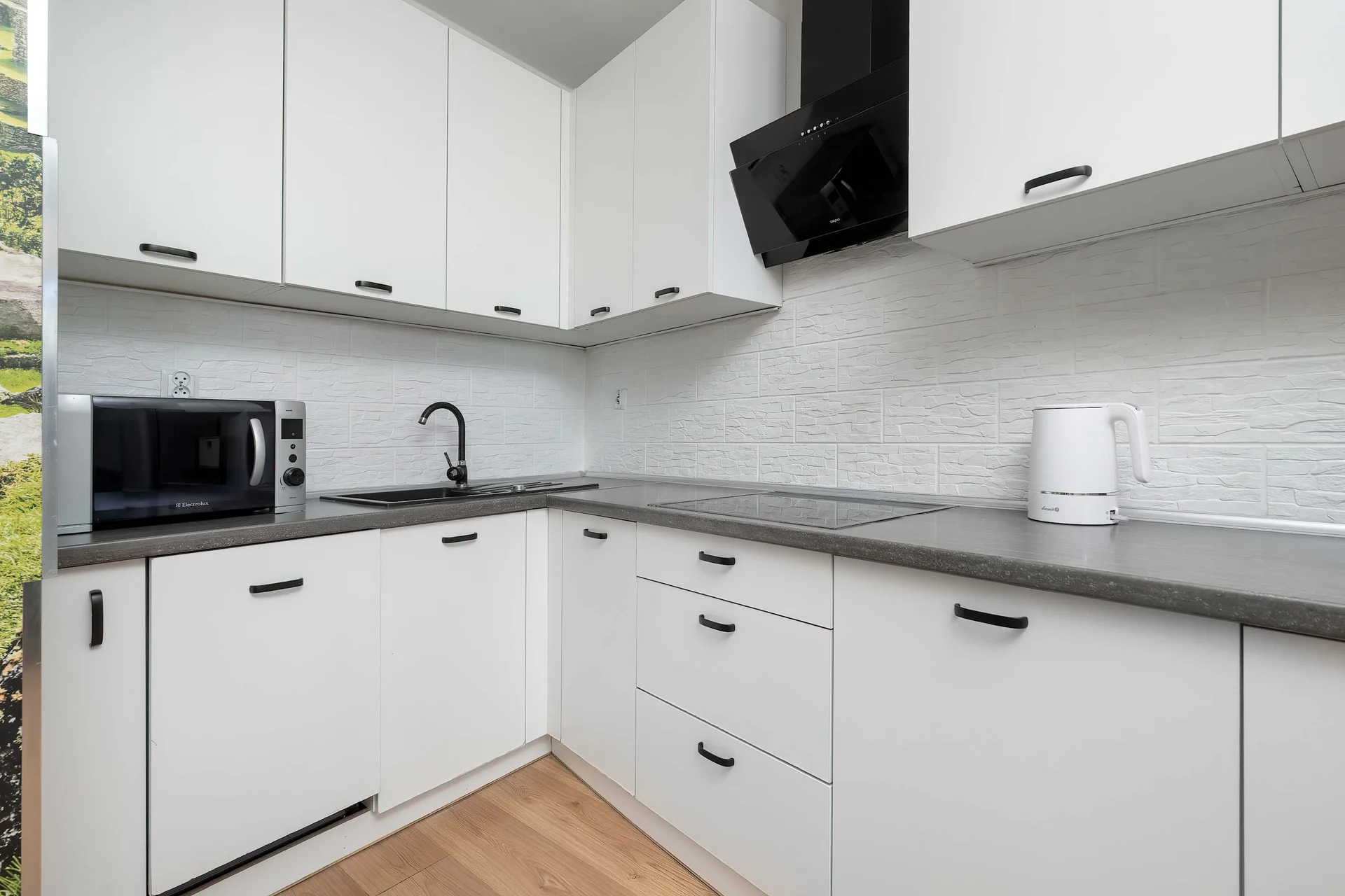 A bright kitchenette is equipped with modern white cabinetry and contrasting dark countertops. A black range hood and a small microwave are neatly arranged in the cooking space.