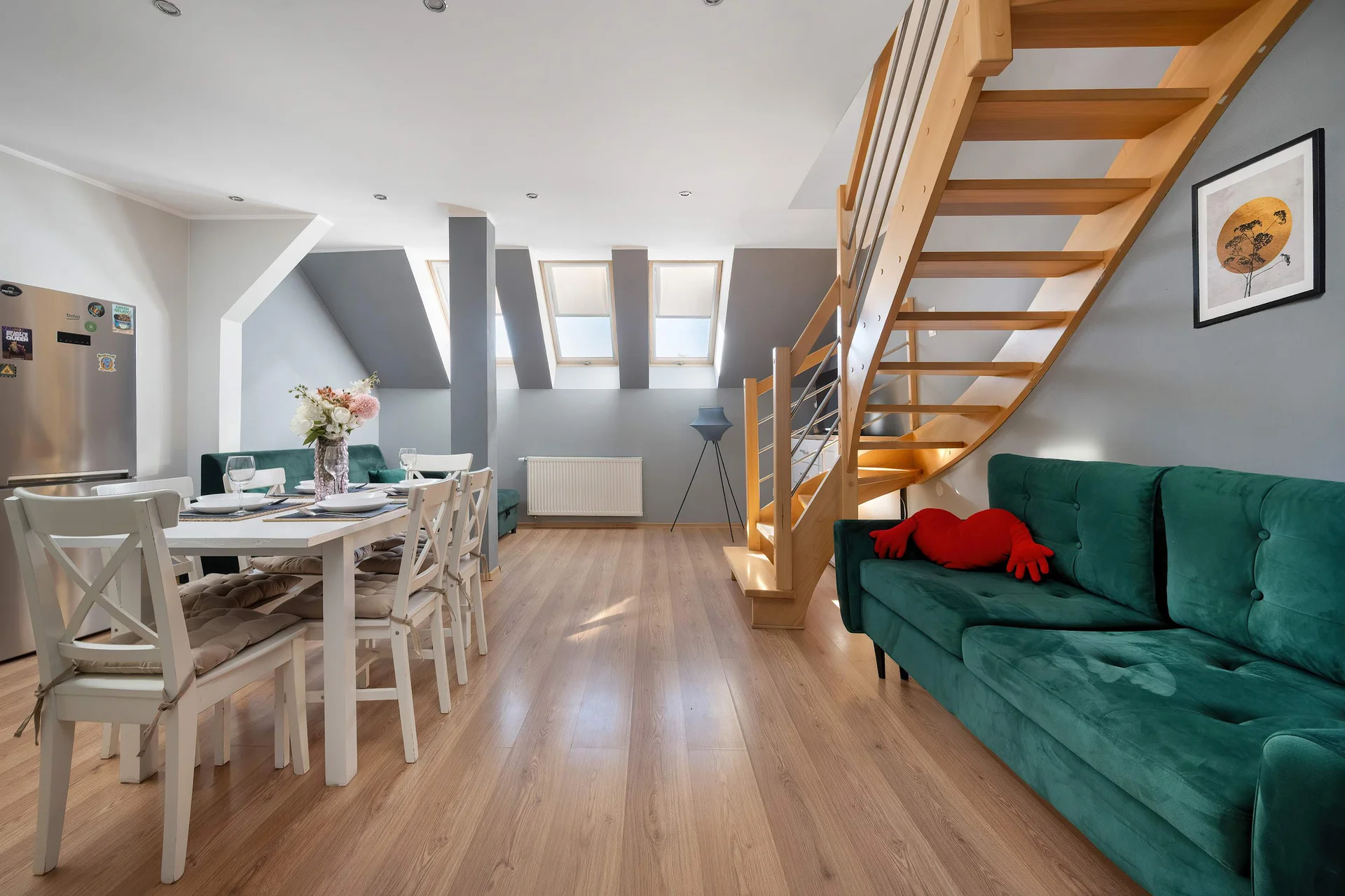 An open-plan living space features a deep green velvet sofa, a wooden staircase, and a dining area. Sunlight filters through the skylights, naturally illuminating the light wooden flooring.
