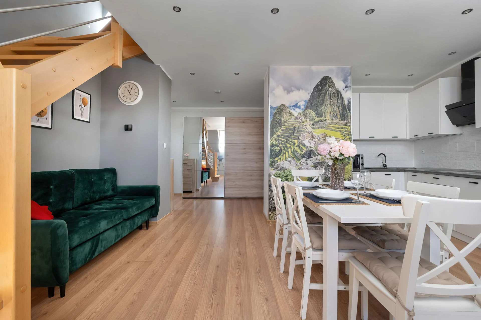 A spacious living and dining room combines a green velvet sofa with a large wooden staircase. A striking mountain landscape mural covers the wall directly behind the dining set.