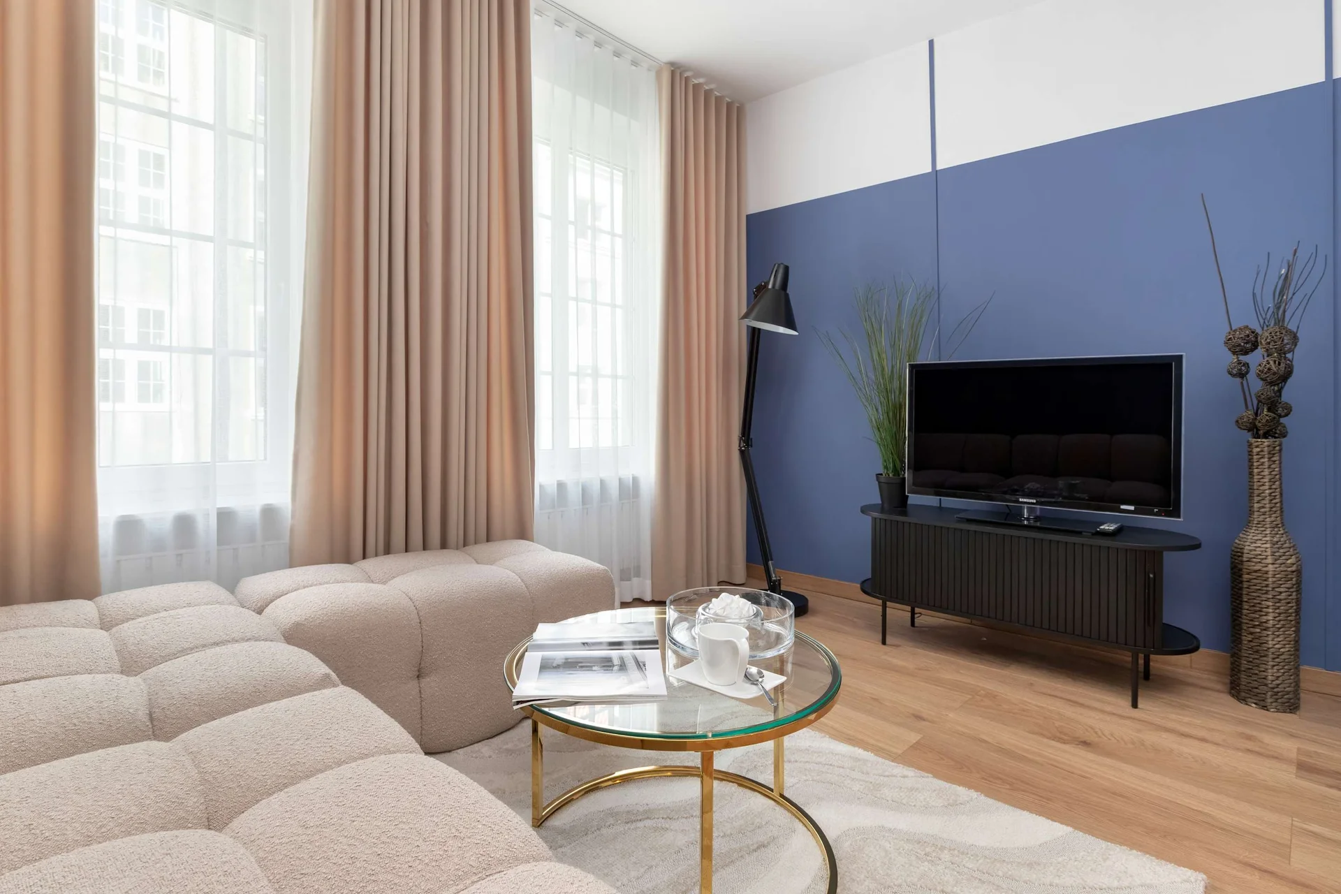 The living space includes a black TV console positioned against a royal blue wall. A plush beige sectional sofa faces a round glass coffee table on light wood floors.