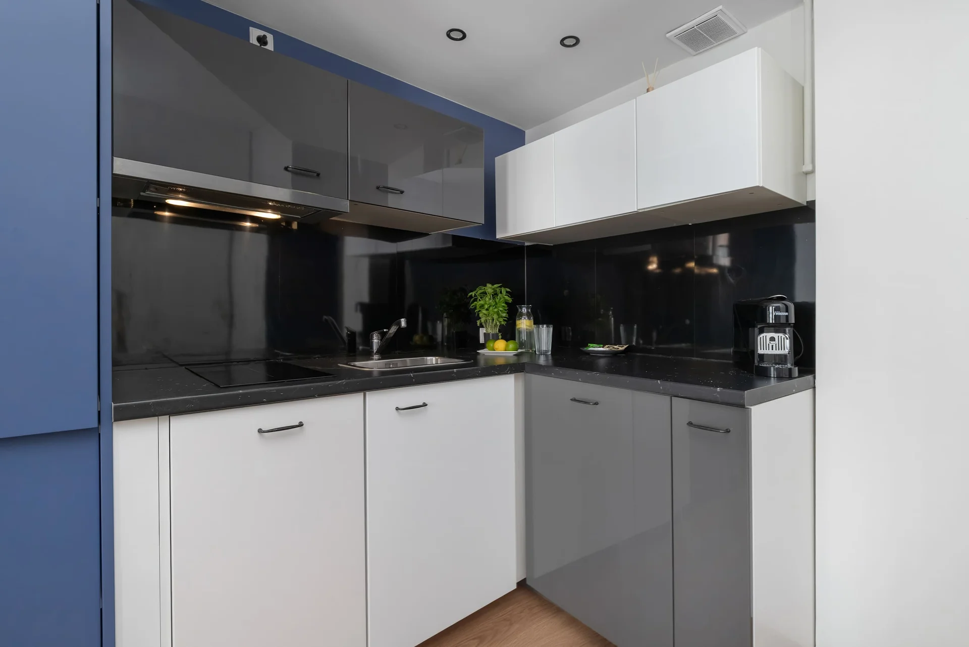 A compact L-shaped kitchen combines sleek white lower cabinets with dark grey upper units. Integrated black appliances fit neatly into the corner.