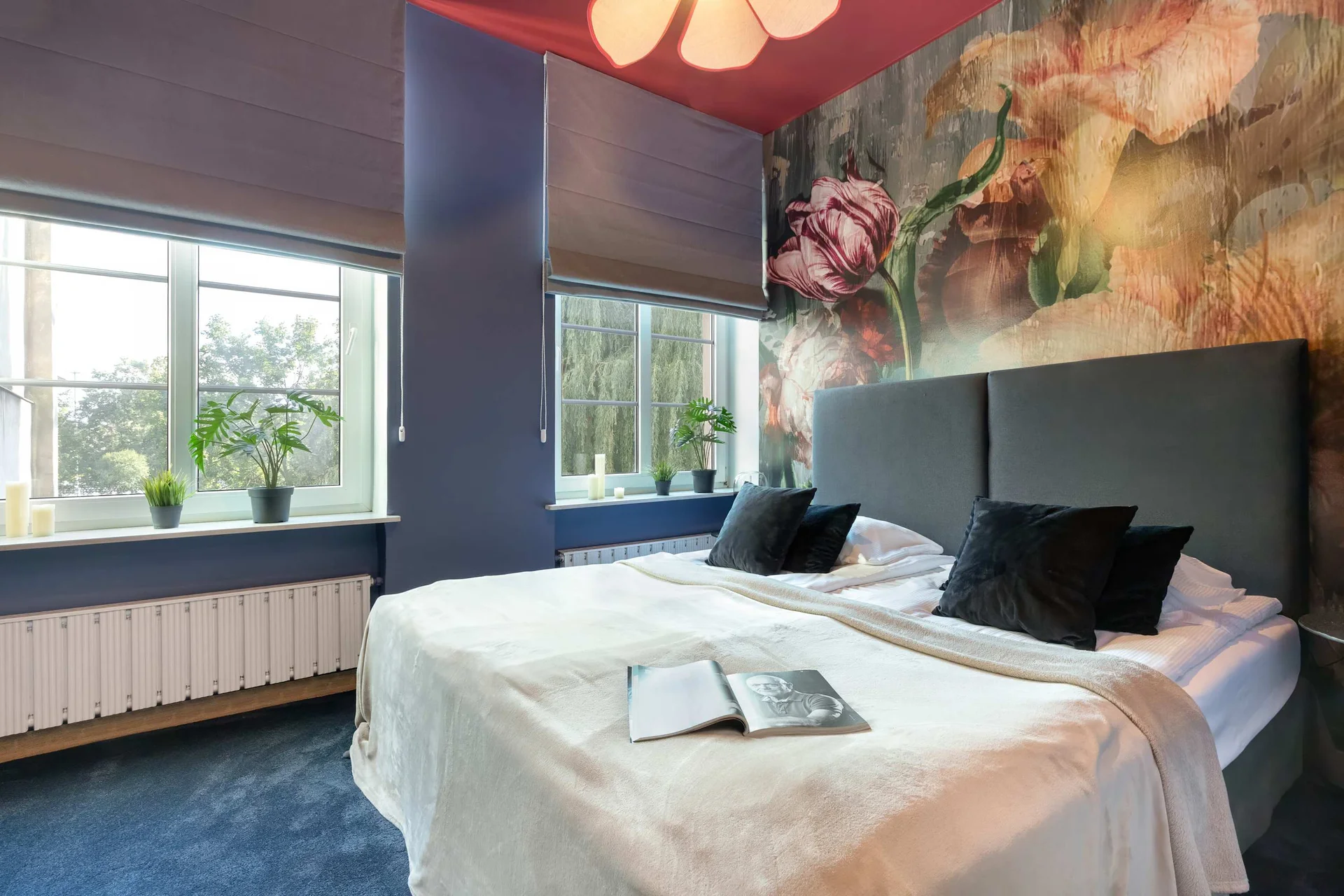 The bedroom showcases a double bed styled with dark velvet pillows against a dramatic floral wall mural. Wide windows flood the blue-walled room with light.