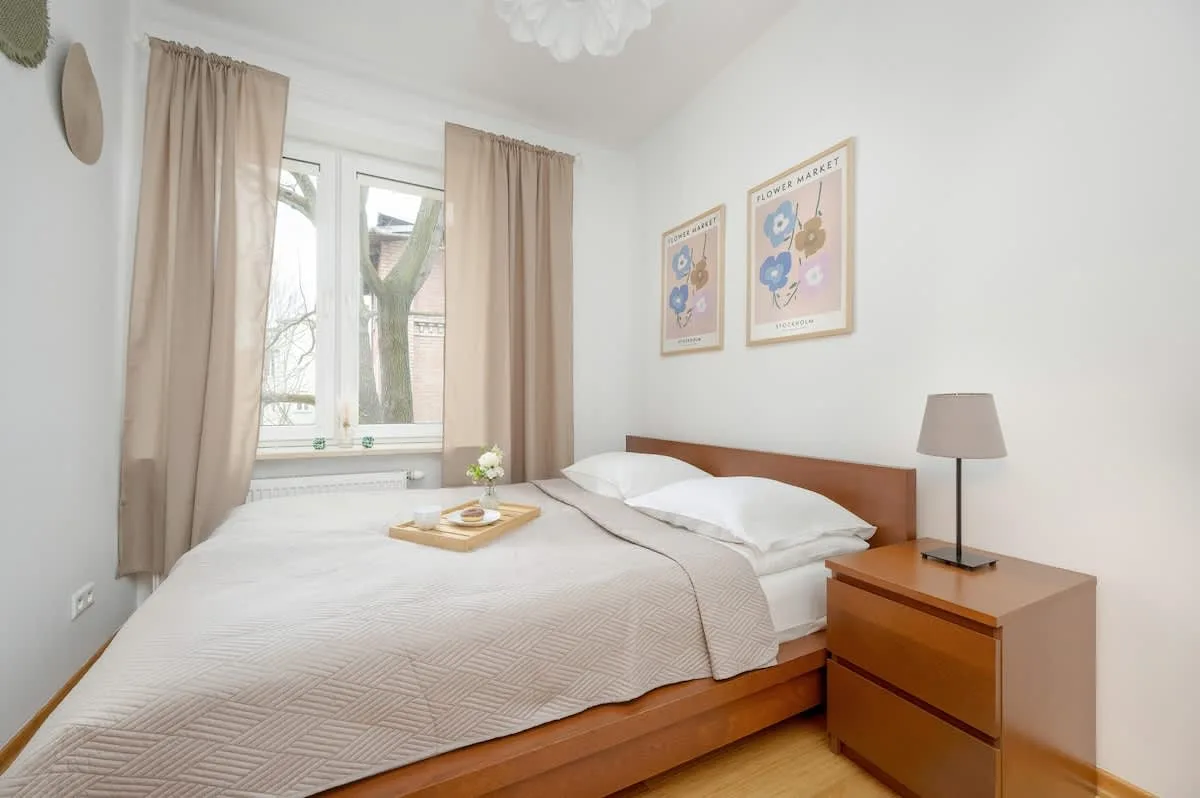 A bright and inviting bedroom featuring a soft white bed, a wooden nightstand, and a large window allowing natural light to enhance the space.