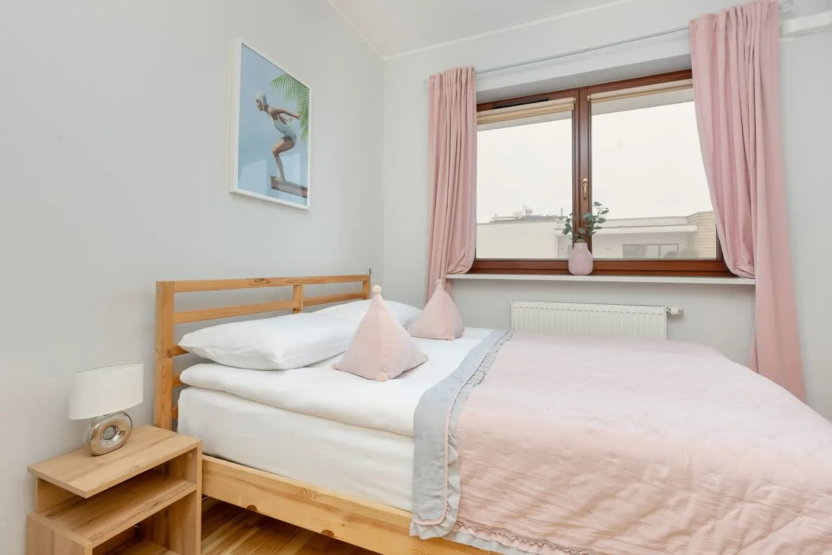 A bright bedroom with a wooden bed, soft pink bedding, and large window, creating a cozy and inviting atmosphere.