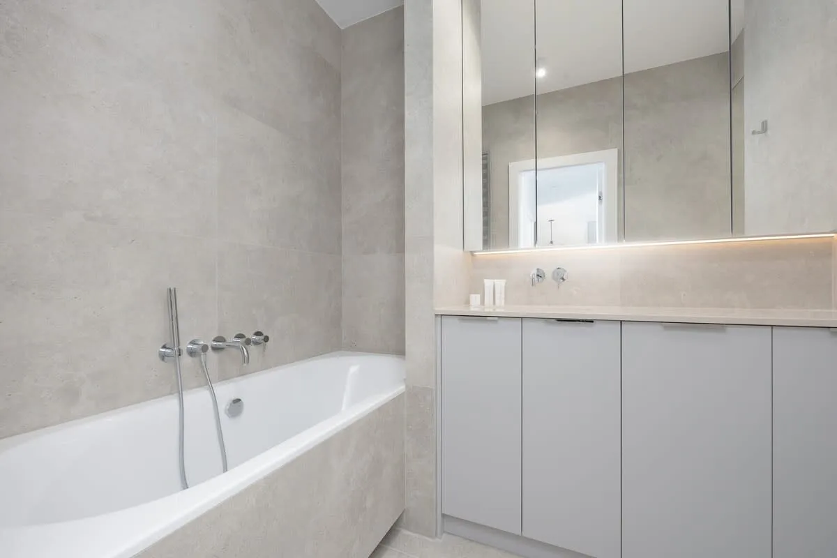A bathroom with a bathtub and overhead shower, grey tiling throughout, and a modern vanity with a mirror.
