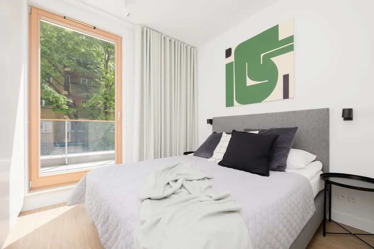 A cozy bedroom with a double bed, large windows, and a green decorative element on the wall above the bed.