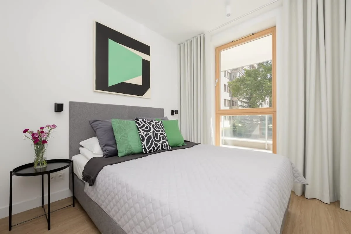A bedroom with a similar setup but featuring a different geometric artwork and a black side table with a vase of flowers.