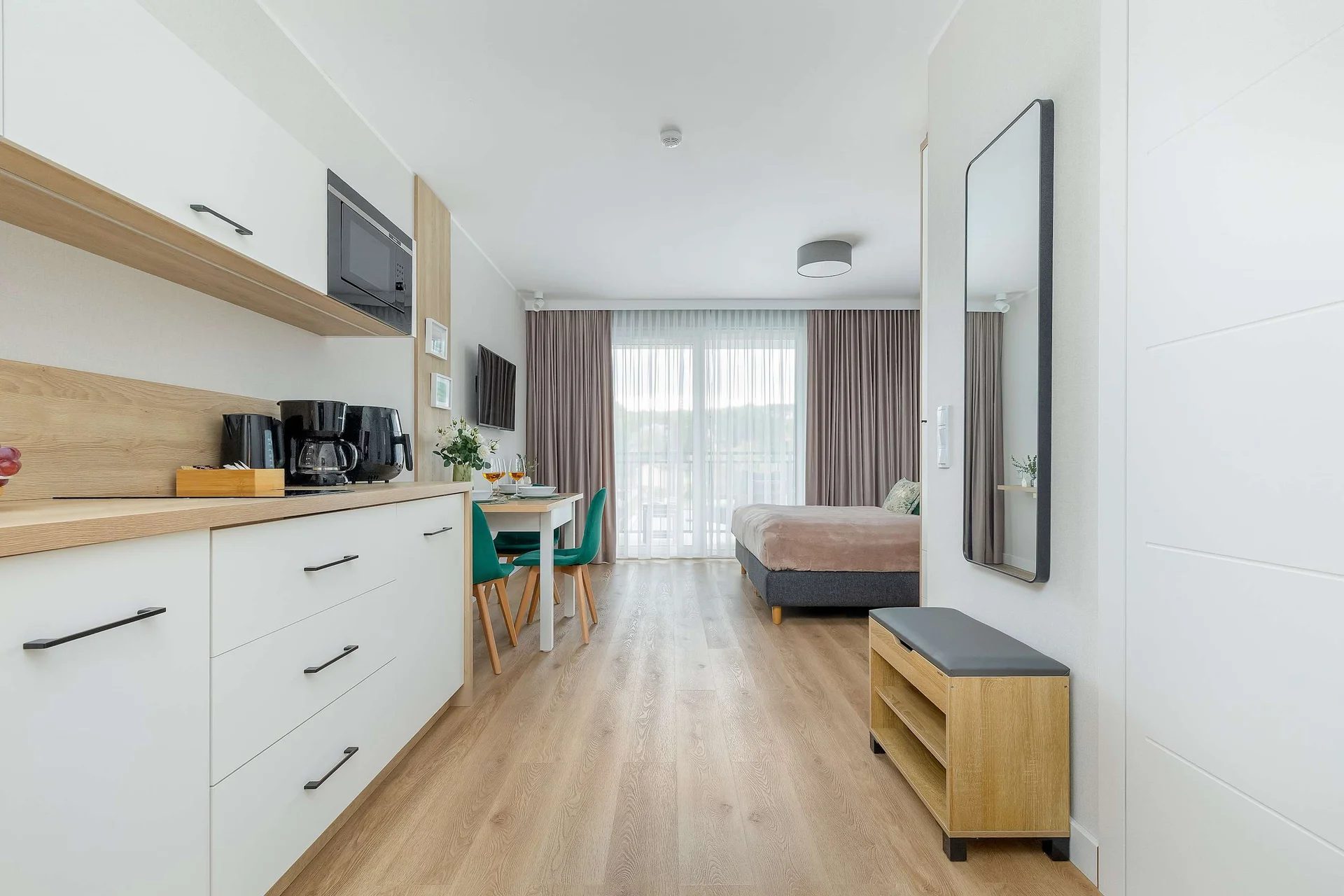 A modern kitchenette with white cabinets, a countertop, and built-in appliances. A wooden floor and a small seating area with a bench and table are visible further into the apartment.