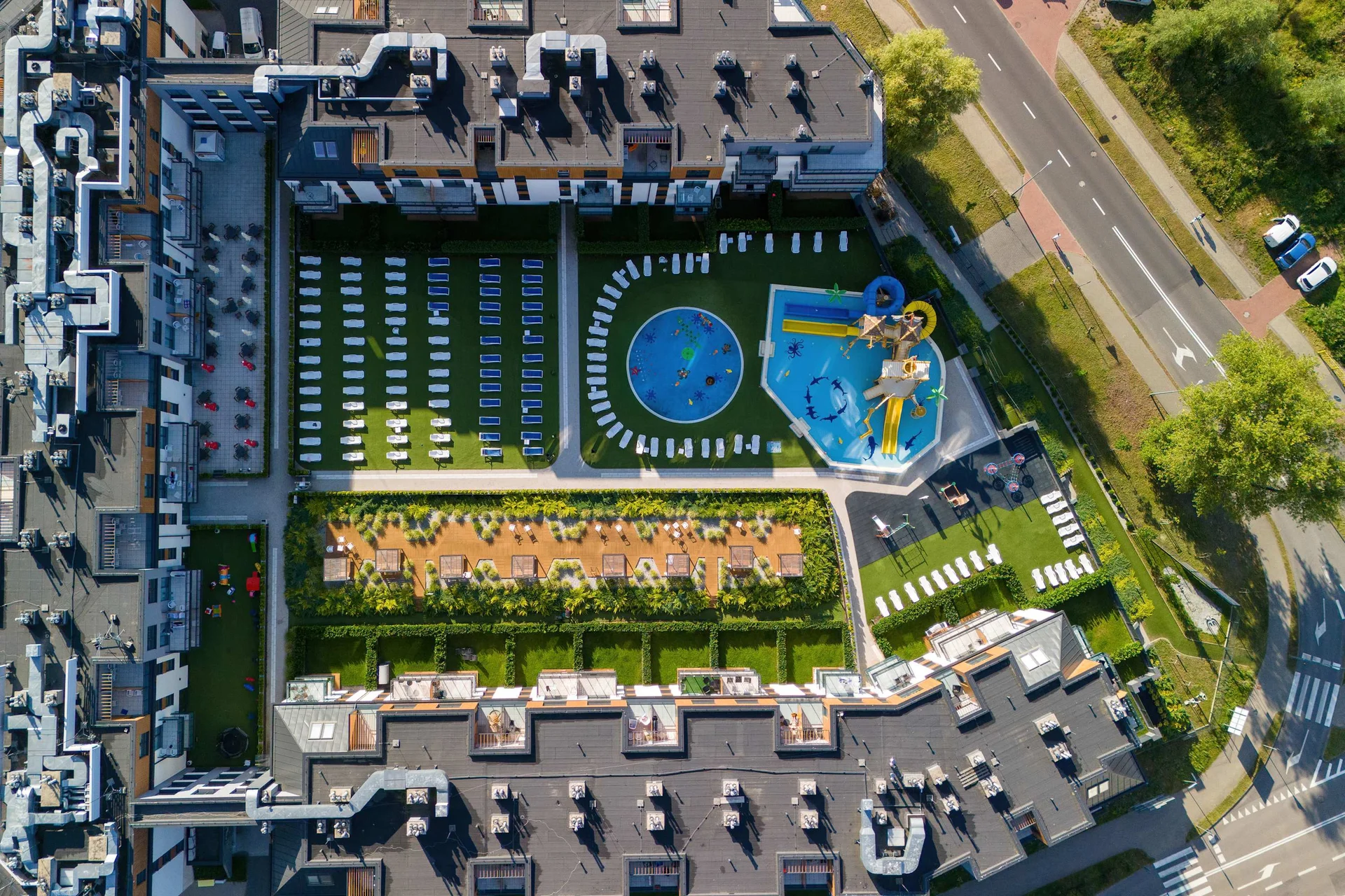 An aerial view of the residential complex showing several modern buildings, surrounding greenery, parking areas, and a blue swimming pool or recreational area in the center.