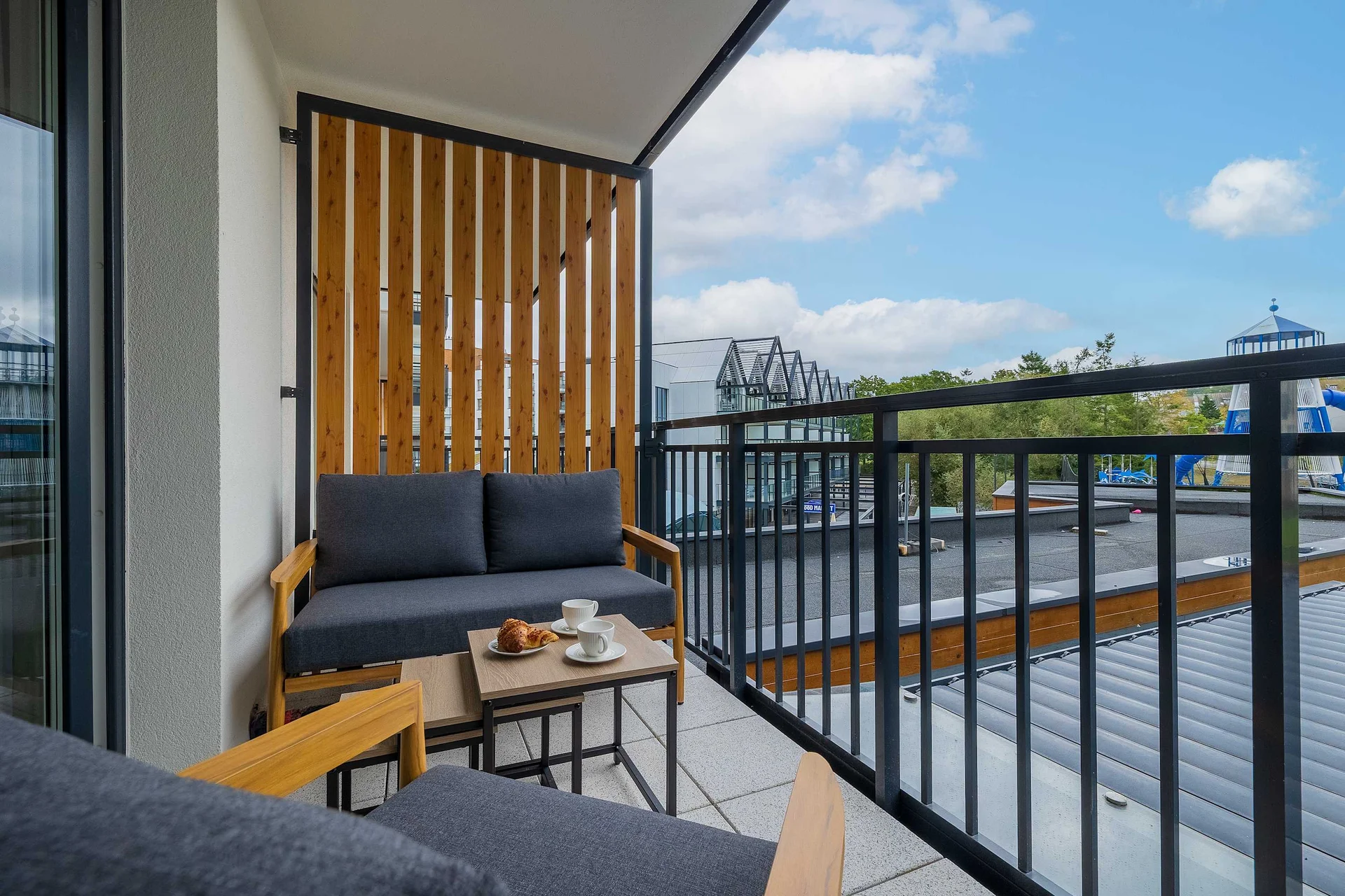 A spacious balcony furnished with comfortable wooden seating and a small table. The balcony offers an open view of the surrounding area and is suitable for relaxing outdoors.