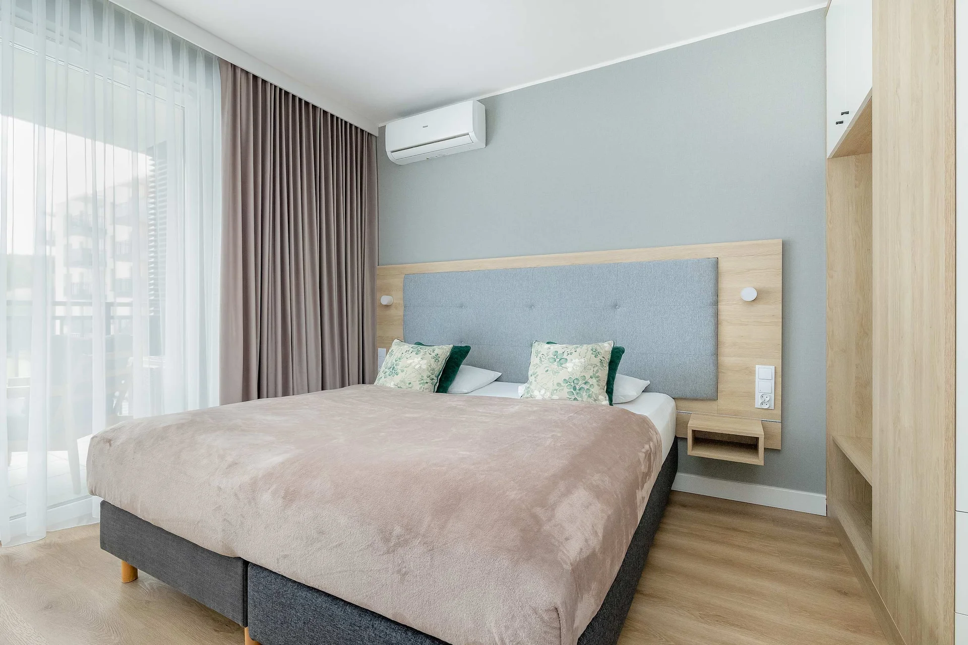 A cozy bedroom featuring a double bed with soft pink bedding and decorative cushions. Large windows with sheer curtains provide plenty of natural light, while a wooden wardrobe stands beside the bed.