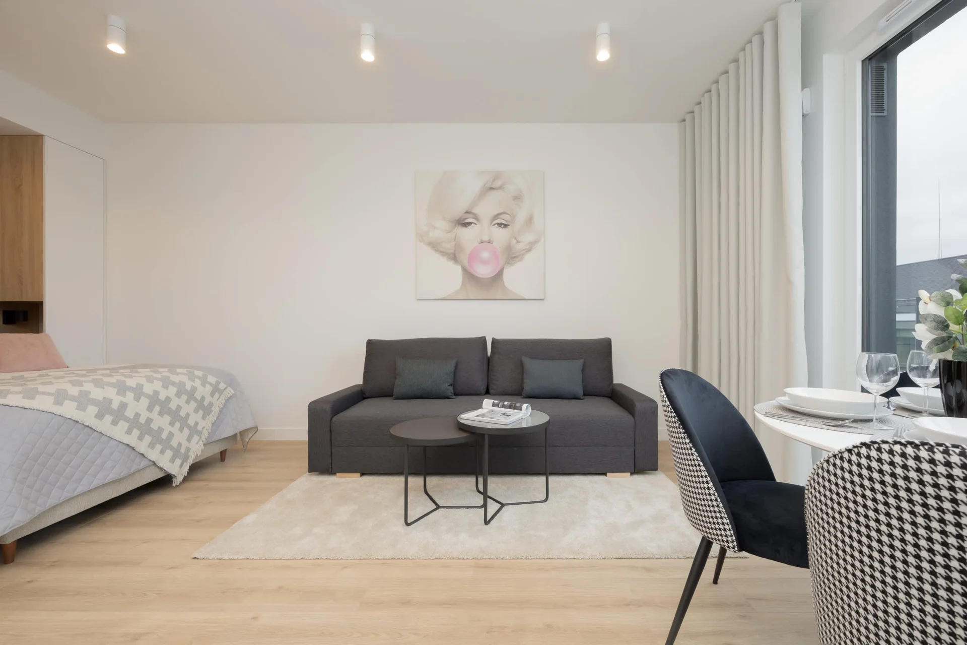 A spacious studio apartment featuring a modern living area with a gray sofa, a round coffee table, and a large wall art of Marilyn Monroe.