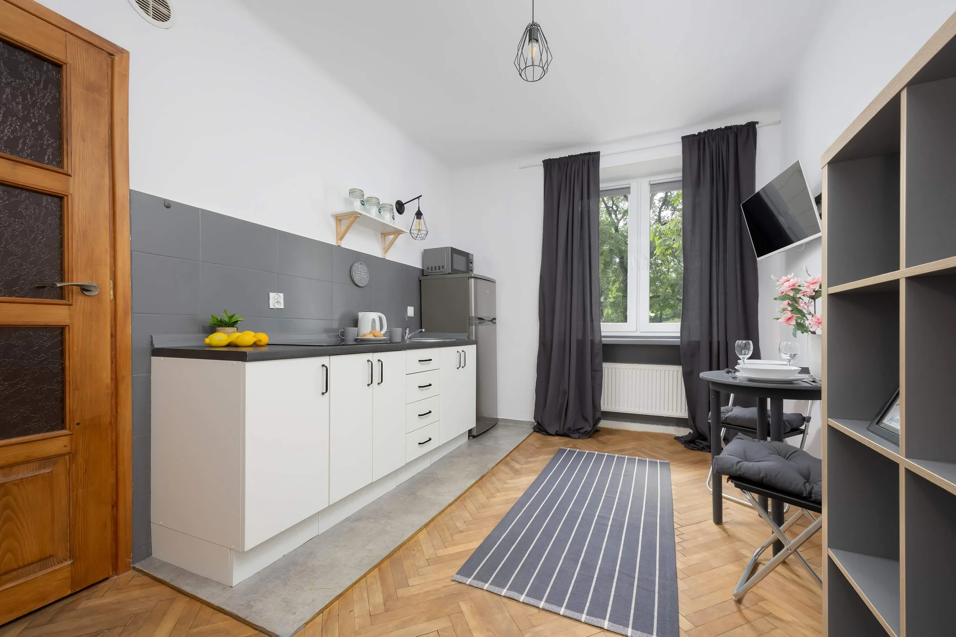 The kitchenette along one wall, featuring white cabinets, a gray countertop, and basic appliances. The wooden door and parquet flooring add warmth to the space.