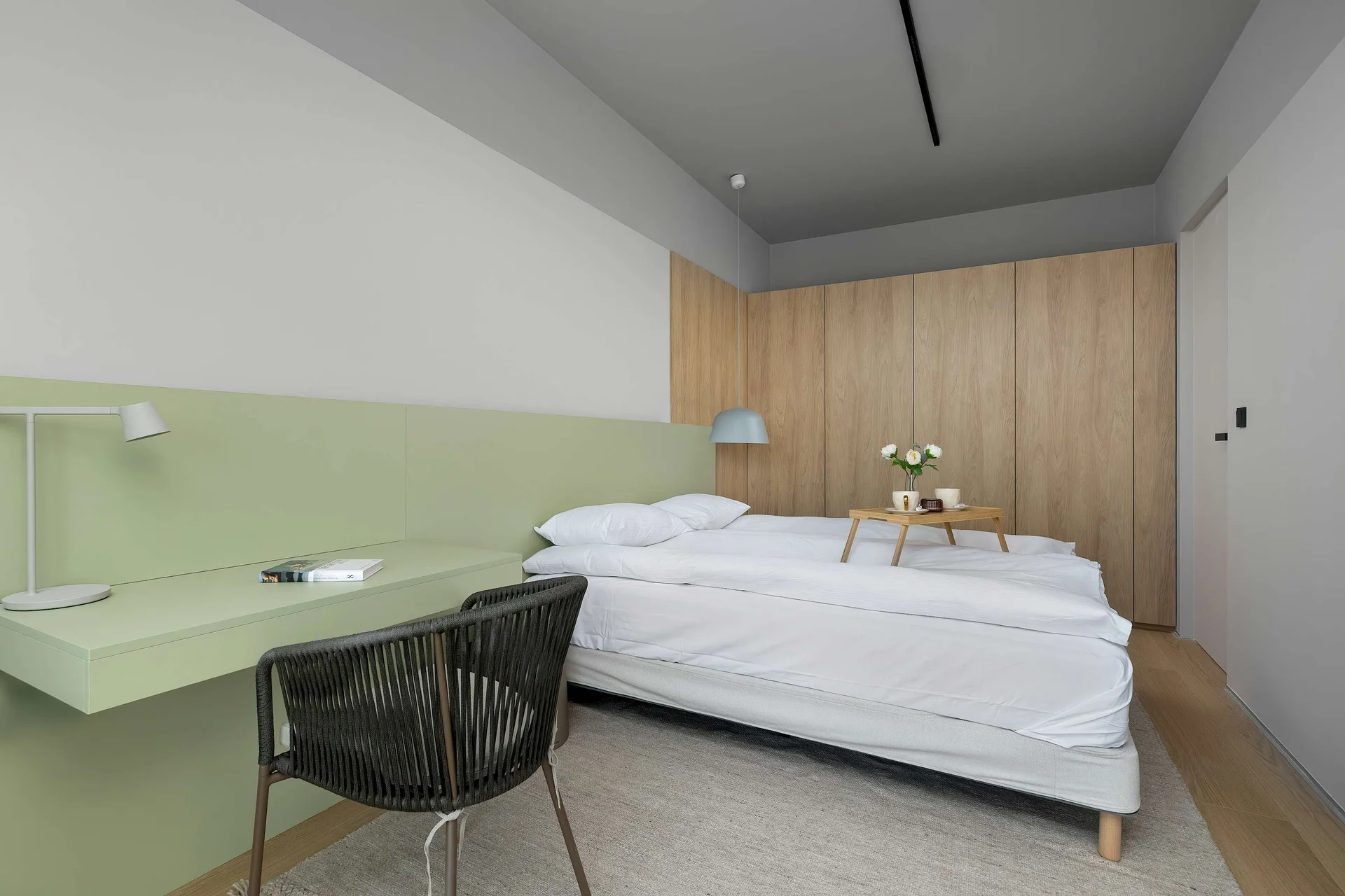 A bedroom with a unique two-tone wall design combining soft green and wood finishes. The bed is centrally placed, creating a clean and balanced composition.