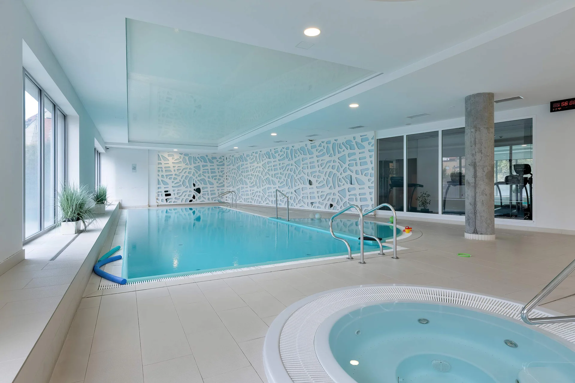 An indoor swimming pool area bathed in natural light from large windows. A round jacuzzi is situated near the main pool, offering a relaxing amenity.