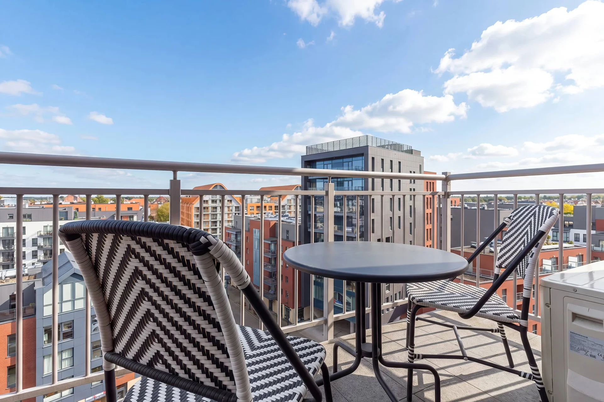 An outdoor balcony setup with a dark wicker chair and a small round table. The elevated viewpoint offers a panoramic view of the surrounding cityscape under a blue sky.