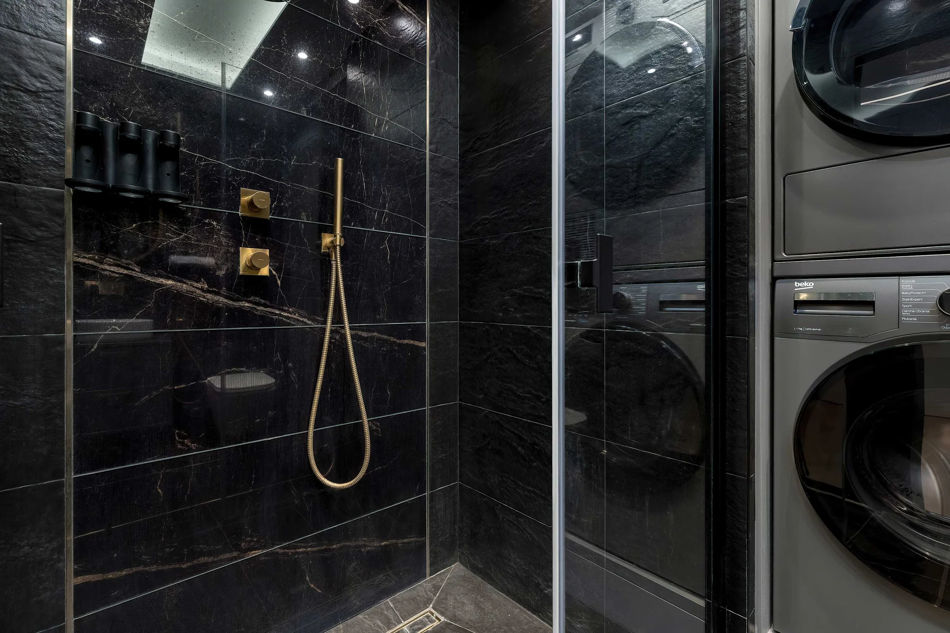 A sleek, modern bathroom featuring black wall tiles and a glass-enclosed shower. A washing machine and dryer are conveniently stacked next to the shower cabin.