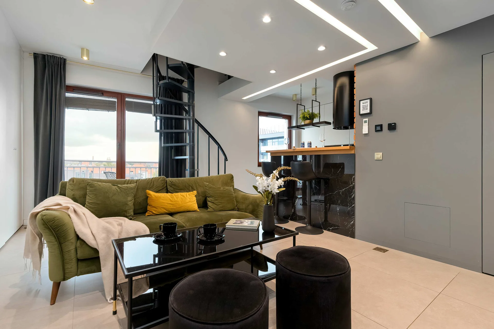 A modern living room featuring a green sofa, black coffee tables, and a spiral staircase. The open-plan design connects to a kitchen area with dark cabinetry.
